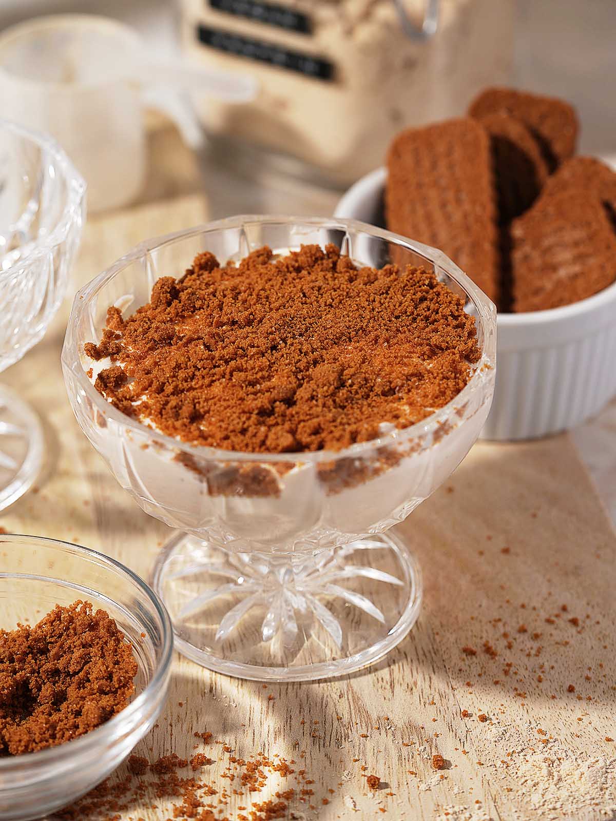 A glass dessert dish filled with Protein Cheesecake and topped with a thick layer of crumbled cookies, with more whole cookies and crumbs nearby on a wooden surface.