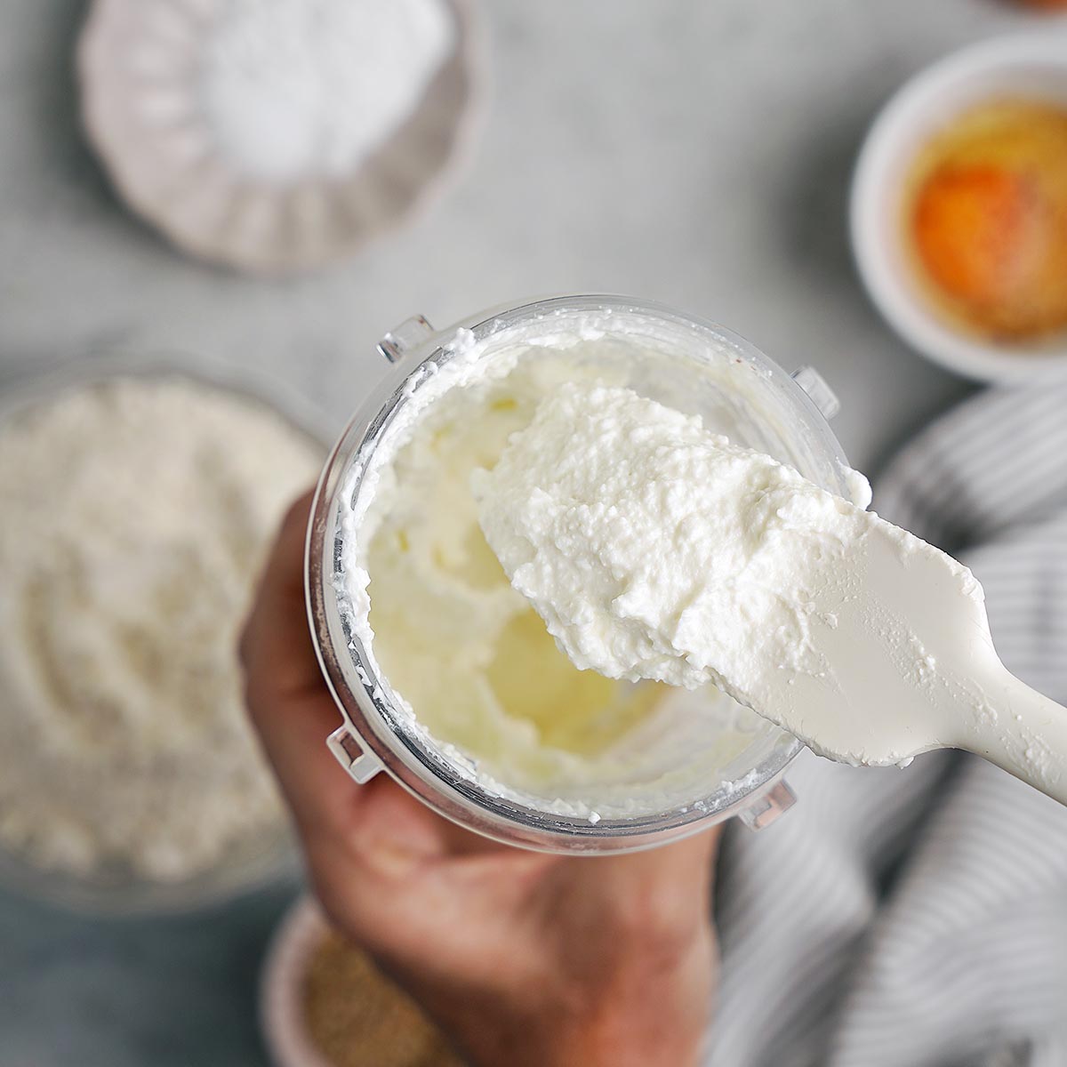 A hand holds a container of whipped ricotta cheese with a spatula, surrounded by bowls of flour, sugar, and an egg on a light surface.