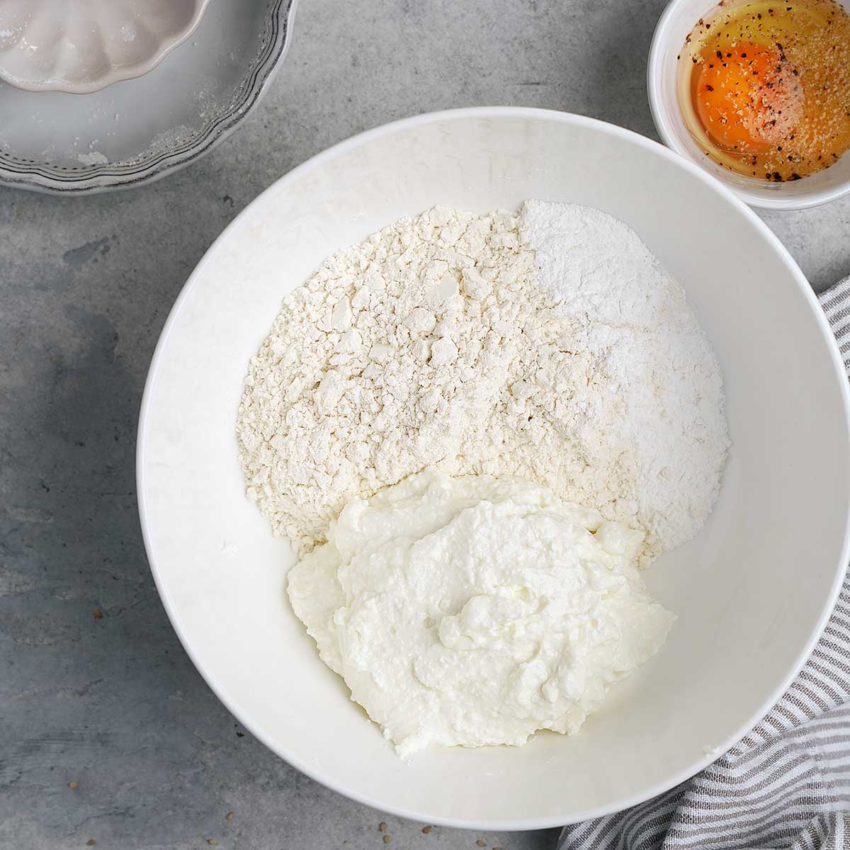 A white bowl with flour and ricotta cheese, next to a small bowl holding a cracked egg with pepper. A striped cloth and part of a ceramic dish are also visible on a gray countertop.