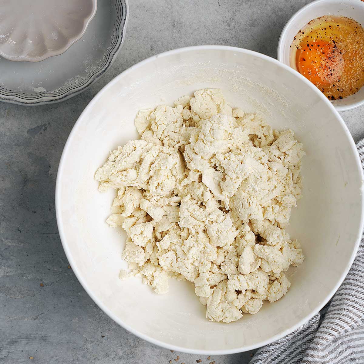 A white bowl with crumbly dough, a small bowl with a cracked egg, salt, and pepper, a striped towel, and a gray plate on a gray countertop.
