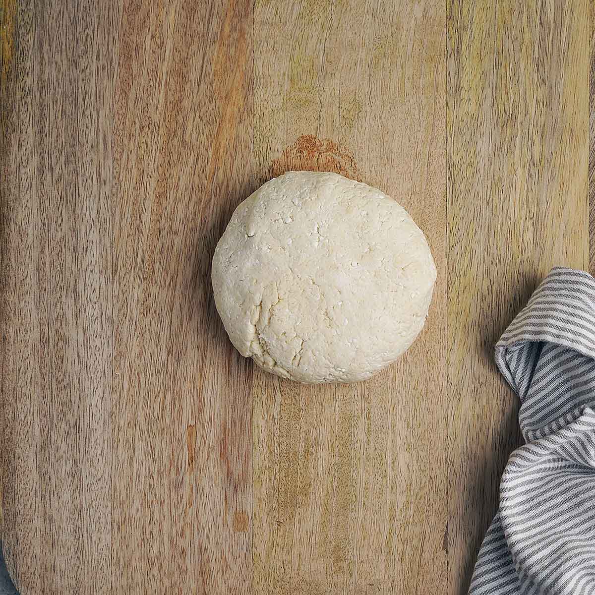 A round ball of dough sits on a wooden surface next to a folded gray and white striped cloth.
