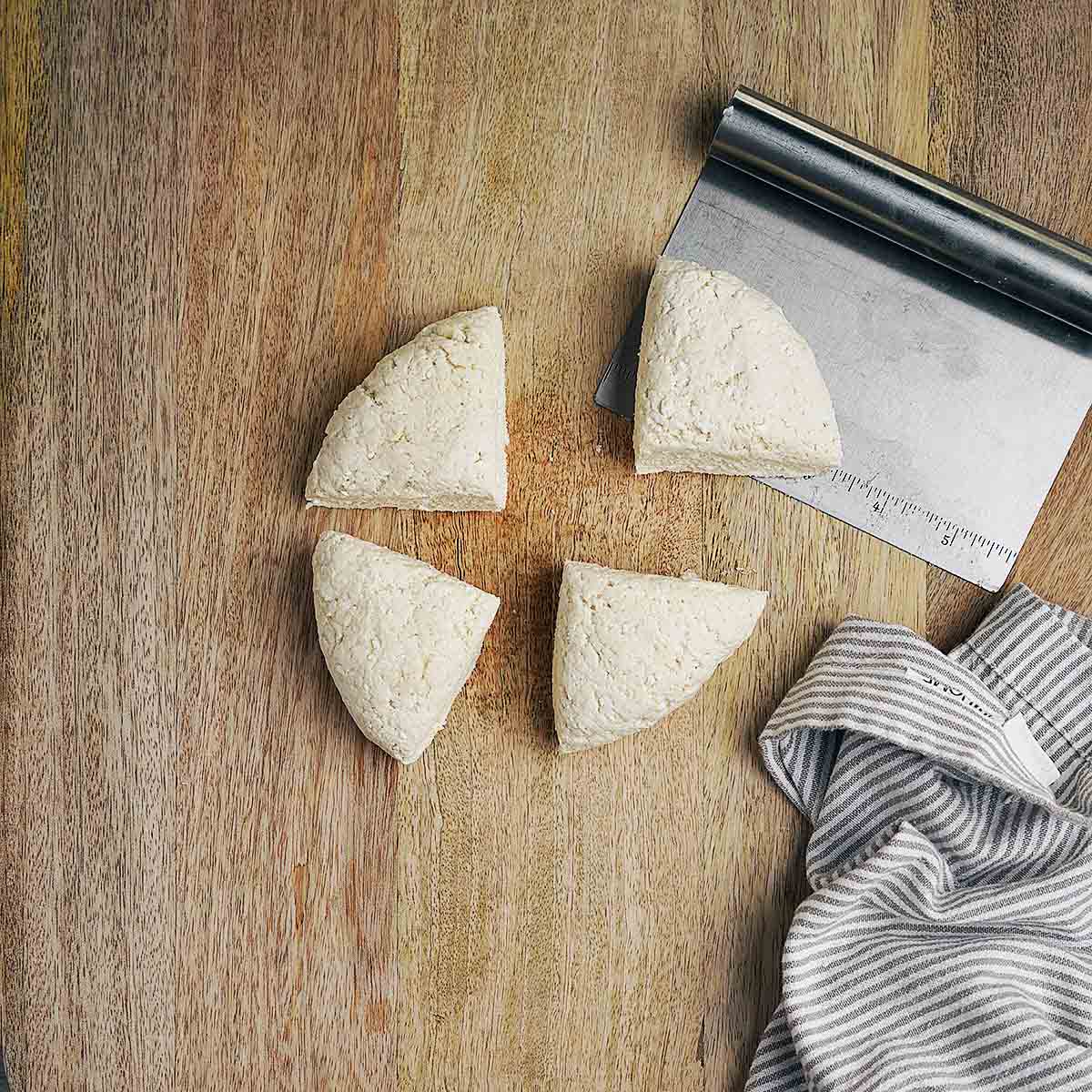 Four dough quarters are arranged in a circle on a wooden surface next to a metal bench scraper and a striped kitchen towel.