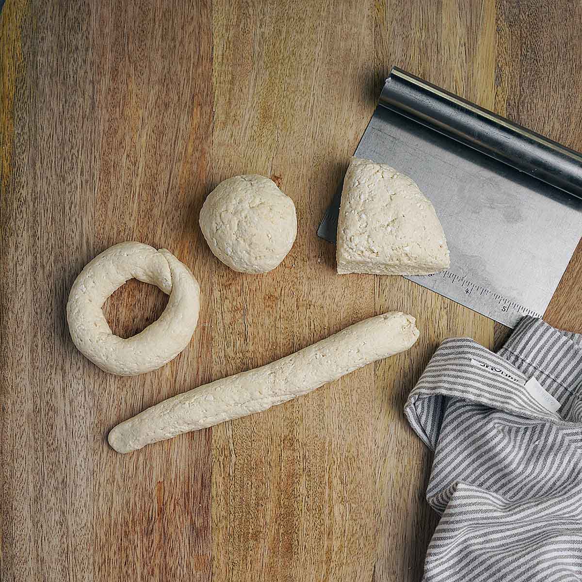 Four pieces of dough in different shapes on a wooden surface, with a metal bench scraper and a striped towel nearby.