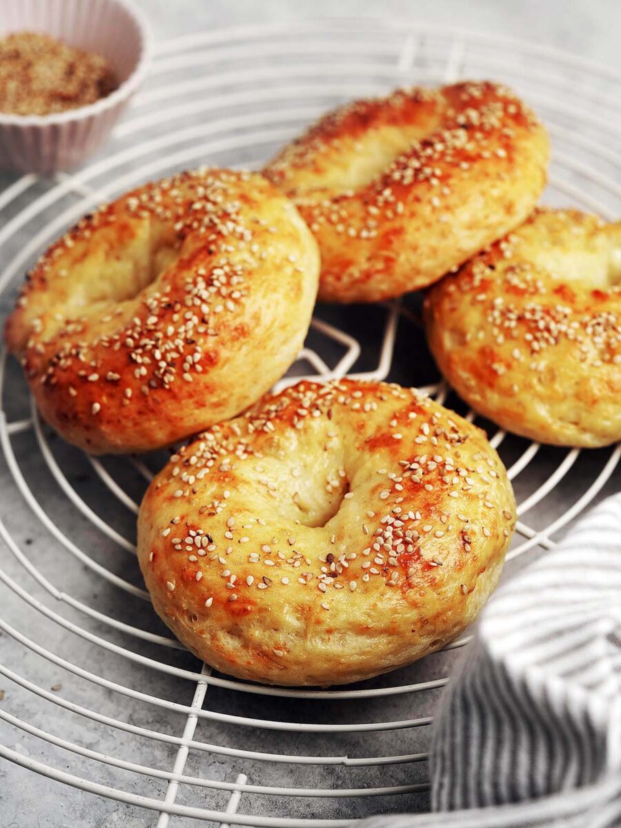 Four golden brown, homemade protein bagels topped with sesame seeds sit on a round wire cooling rack, with a small bowl of sesame seeds and a striped cloth nearby.