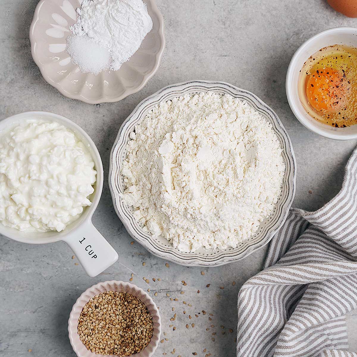 Bowl of flour, egg with pepper in a dish, baking powder, cottage cheese in a cup, sesame seeds, and a striped towel on a gray surface.
