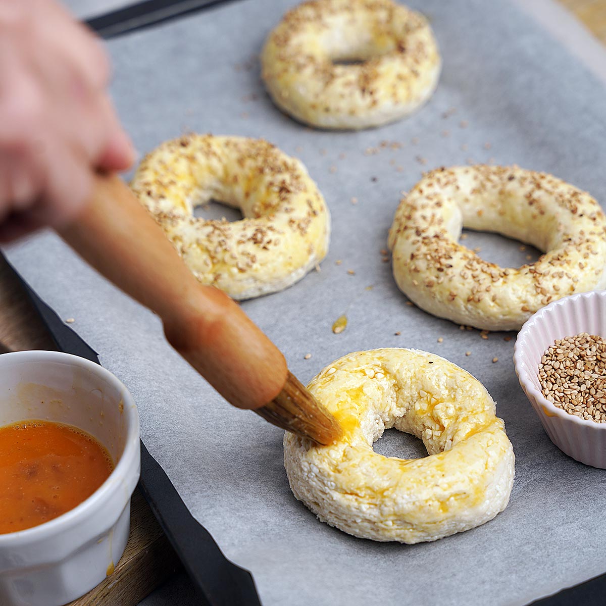 A hand brushes egg wash onto an unbaked ring-shaped dough on a baking tray with parchment paper. Three other dough rings and a small bowl of sesame seeds are nearby.