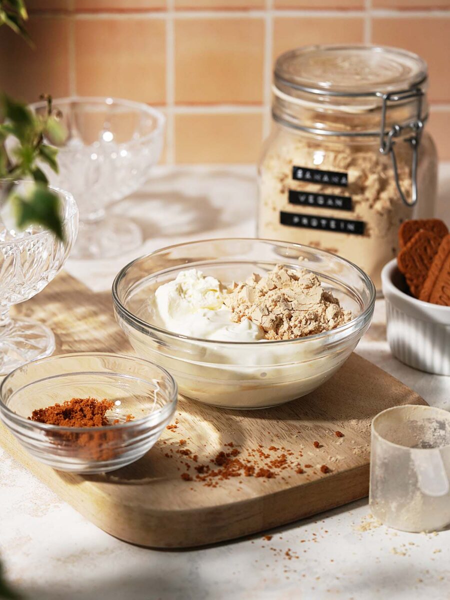 A glass bowl with yogurt and protein powder on a wooden board, a smaller bowl with cocoa powder, a jar labeled VEGAN PROTEIN, and a dish of cookies, all on a bright kitchen counter.