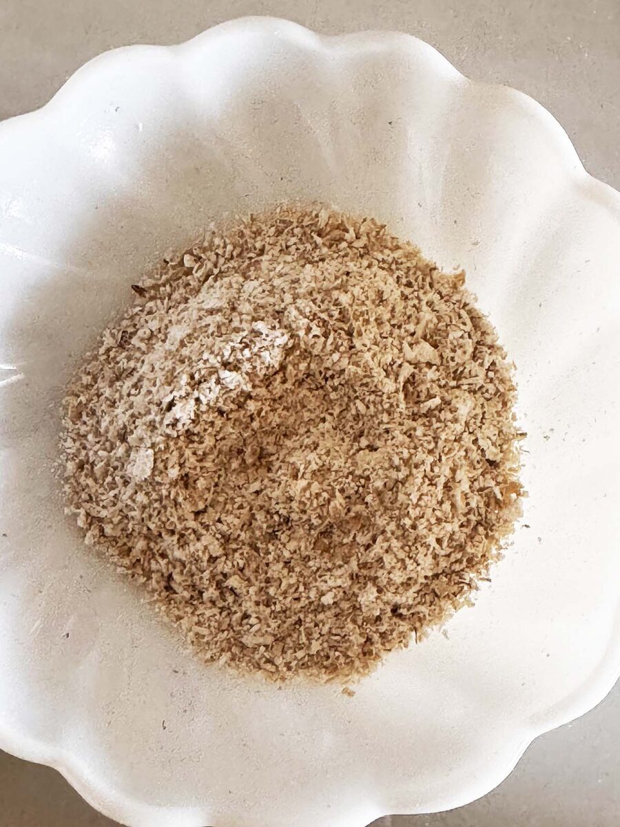 A white scalloped bowl filled with light brown, fine breadcrumbs sits on a beige surface.