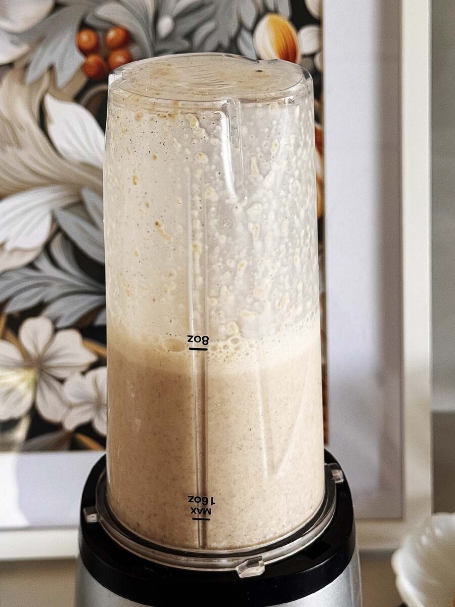 A clear blender cup filled with a creamy smoothie mixture is placed upside down on its blender base. The background features a floral artwork with white and orange flowers.