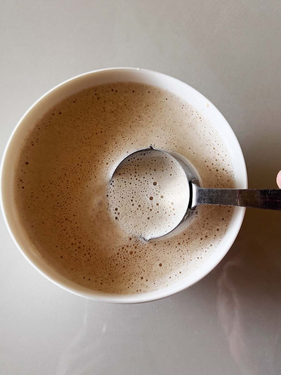 A hand holds a spoon, scooping frothy coffee from a white cup on a light gray surface.