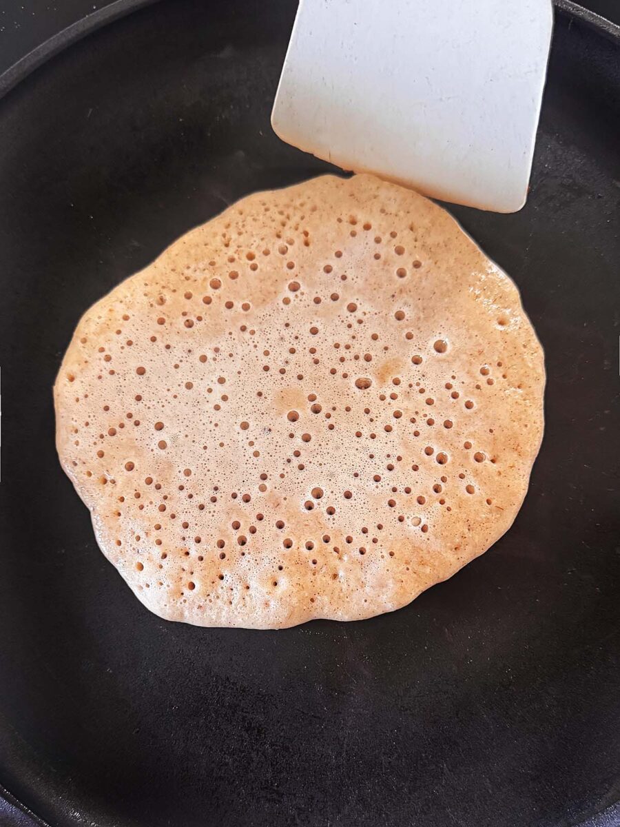 A pancake with bubbles forming on its surface cooks on a black pan, with a white spatula positioned next to it, ready to flip.