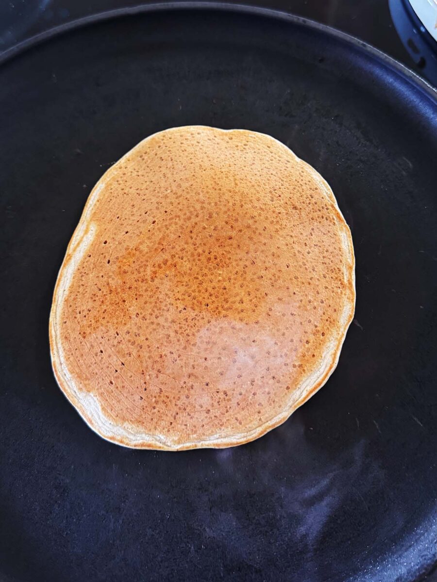 A single golden-brown pancake cooking on a black skillet, viewed from above.