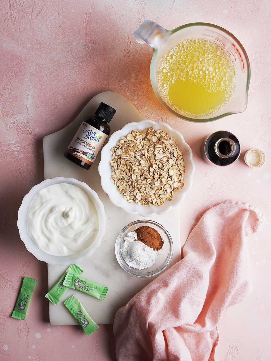 A marble board with oats, yogurt, melted butter, vanilla, sweetener packets, baking powder, and cinnamon on a pink surface beside a pink cloth.