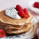 A stack of pancakes topped with whipped cream, chia seeds, raspberries, and edible flowers sits on a decorative plate.