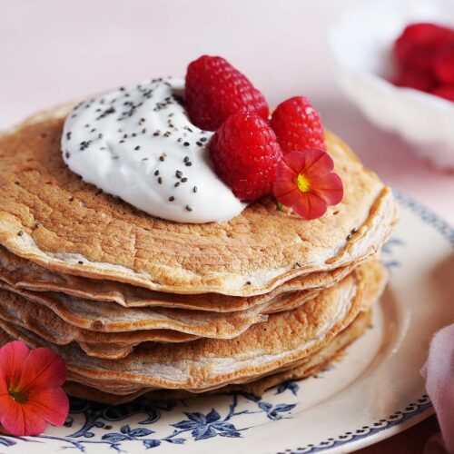 A stack of pancakes topped with whipped cream, chia seeds, raspberries, and edible flowers sits on a decorative plate.