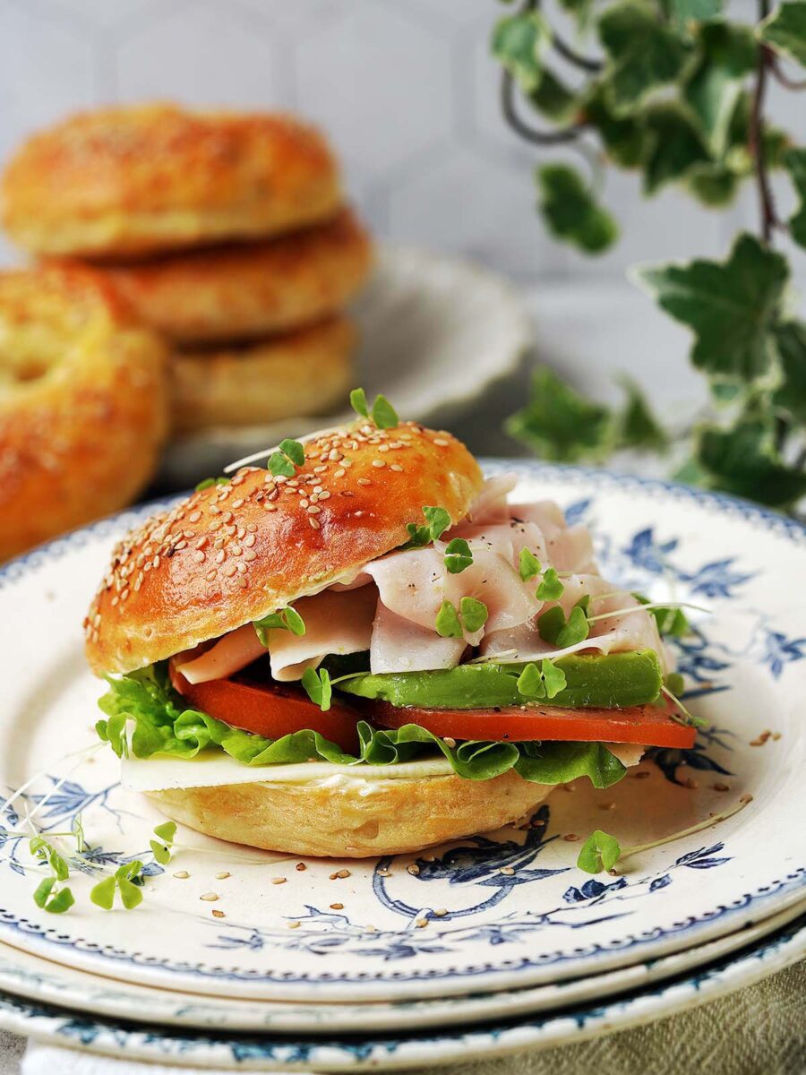 A protein bagel sandwich filled with lettuce, tomato, cucumber, and sliced deli meat, topped with microgreens, sits on a decorative plate. More bagels and green ivy are blurred in the background.