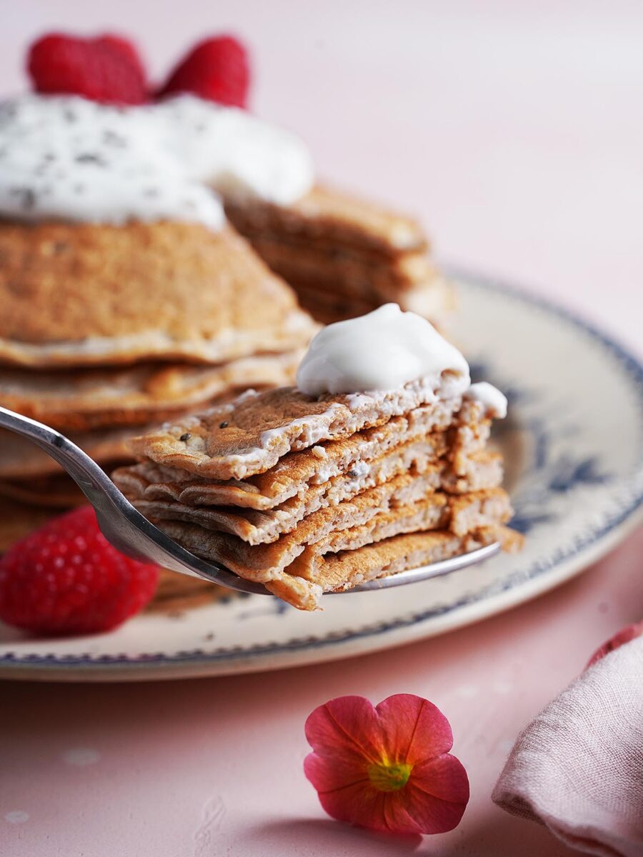 A close-up of a fork holding a bite of fluffy Protein Pancakes Without Protein Powder topped with cream, with a stack of pancakes, raspberries, and a pink flower on a plate in the background.