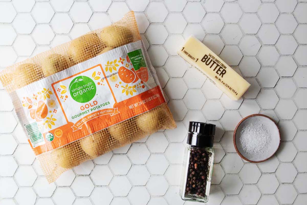 A bag of organic gold potatoes, a stick of butter, a small bowl of salt, and a pepper grinder are arranged on a white hexagonal tile surface.
