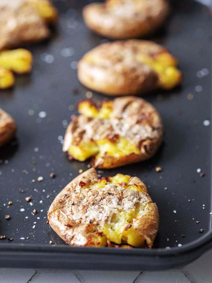 A baking tray with several roasted smashed potatoes sprinkled with salt and pepper, showing crispy, golden edges and fluffy yellow insides.
