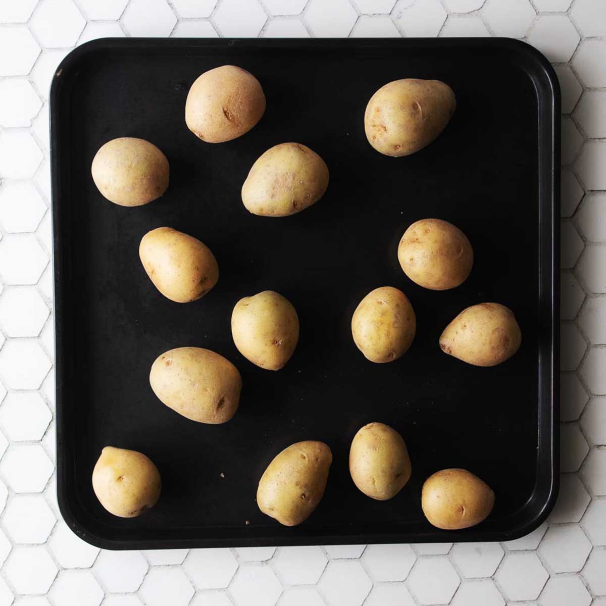 A black baking tray with 14 small, whole potatoes evenly spaced on it, set on a white hexagon-tiled surface.