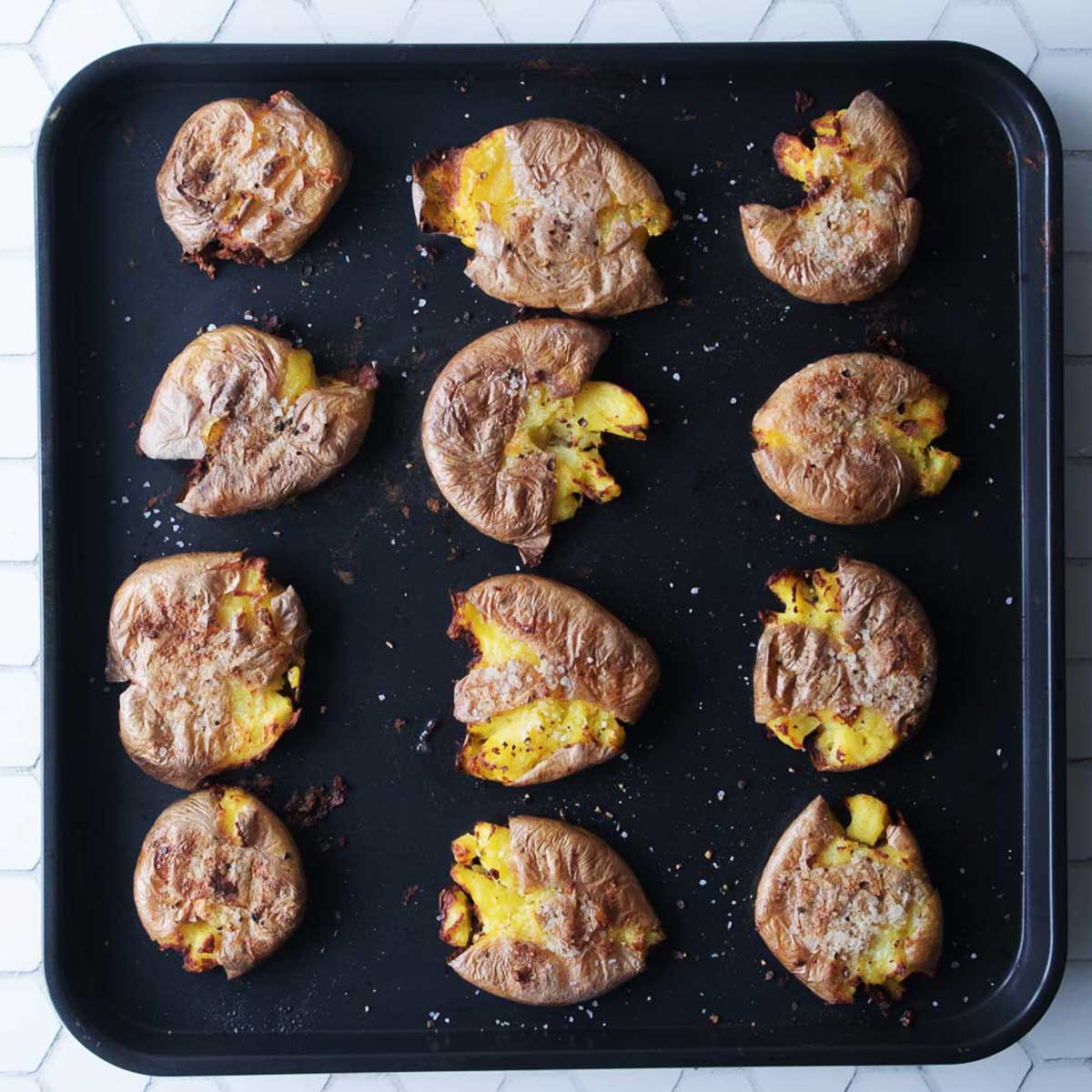 Twelve smashed and roasted potatoes are spread out on a black baking tray, sprinkled with salt and pepper, on a white hexagon-tiled surface.