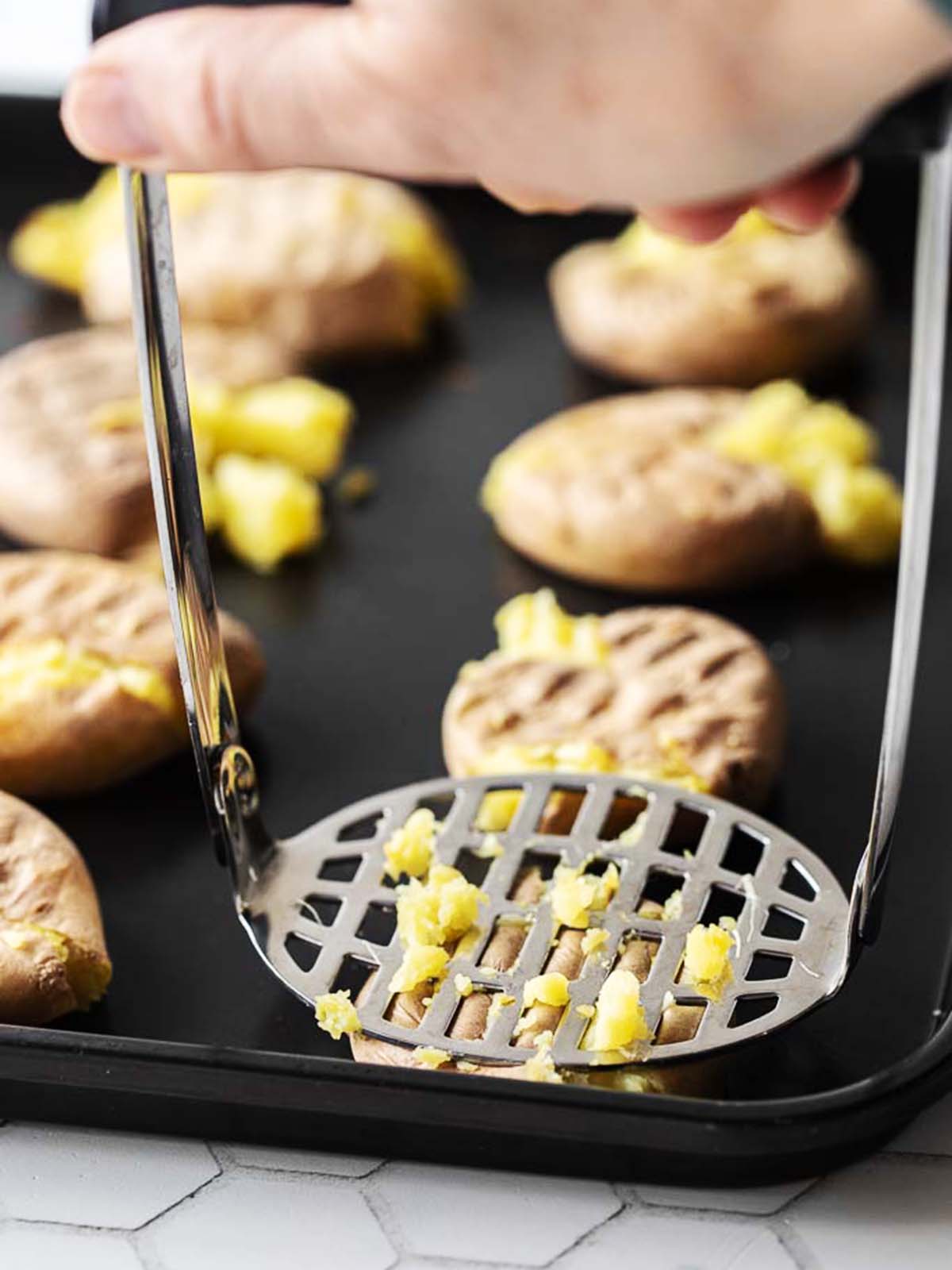 A hand presses a metal masher onto small cooked potatoes on a baking tray, flattening them. Some potatoes are already smashed and spaced out on the tray.