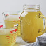 Two glass measuring cups and a mason jar filled with golden-colored broth.