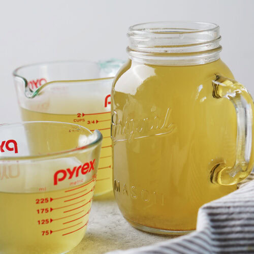Two glass measuring cups and a mason jar filled with golden-colored broth.