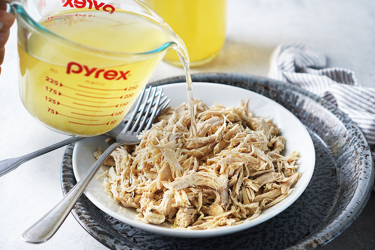 Chicken broth being poured from a measuring cup over a plate of shredded chicken.