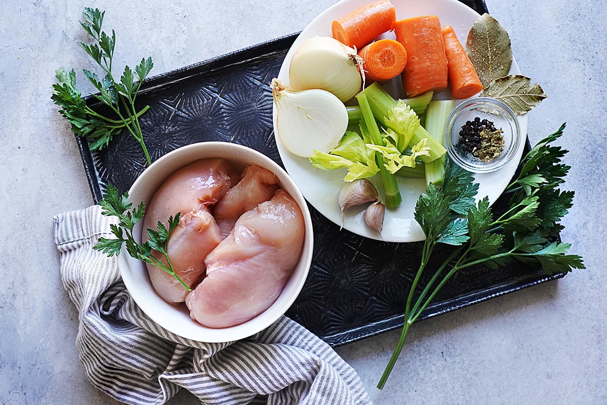 A bowl of raw chicken breasts next to a plate of chopped carrots, onions, celery, garlic, bay leaves, and peppercorns on a black tray.