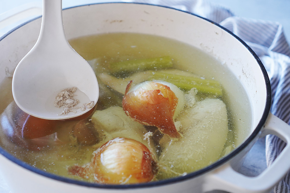 A white ladle lifts foam from a pot of chicken stock containing whole onions and celery stalks.