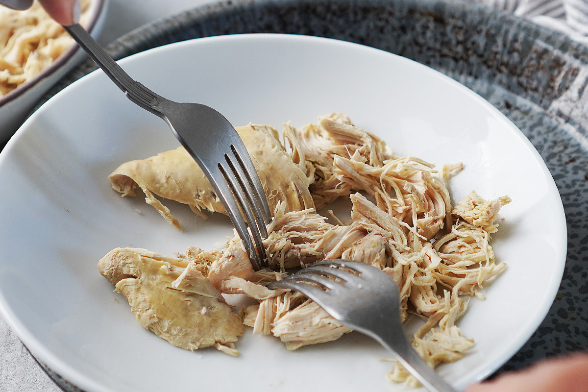 Two forks shredding cooked chicken in a white bowl.