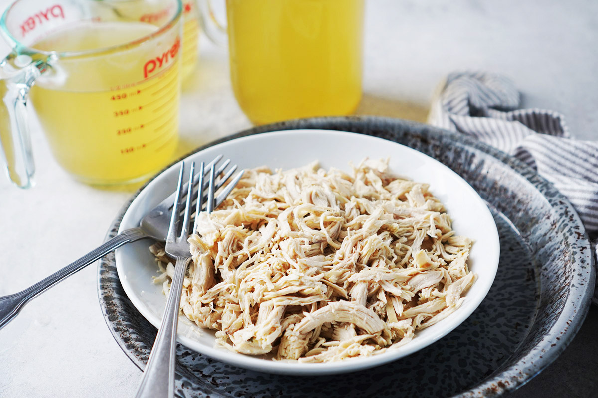 A bowl of shredded chicken with a fork, with measuring cups of broth in the background.