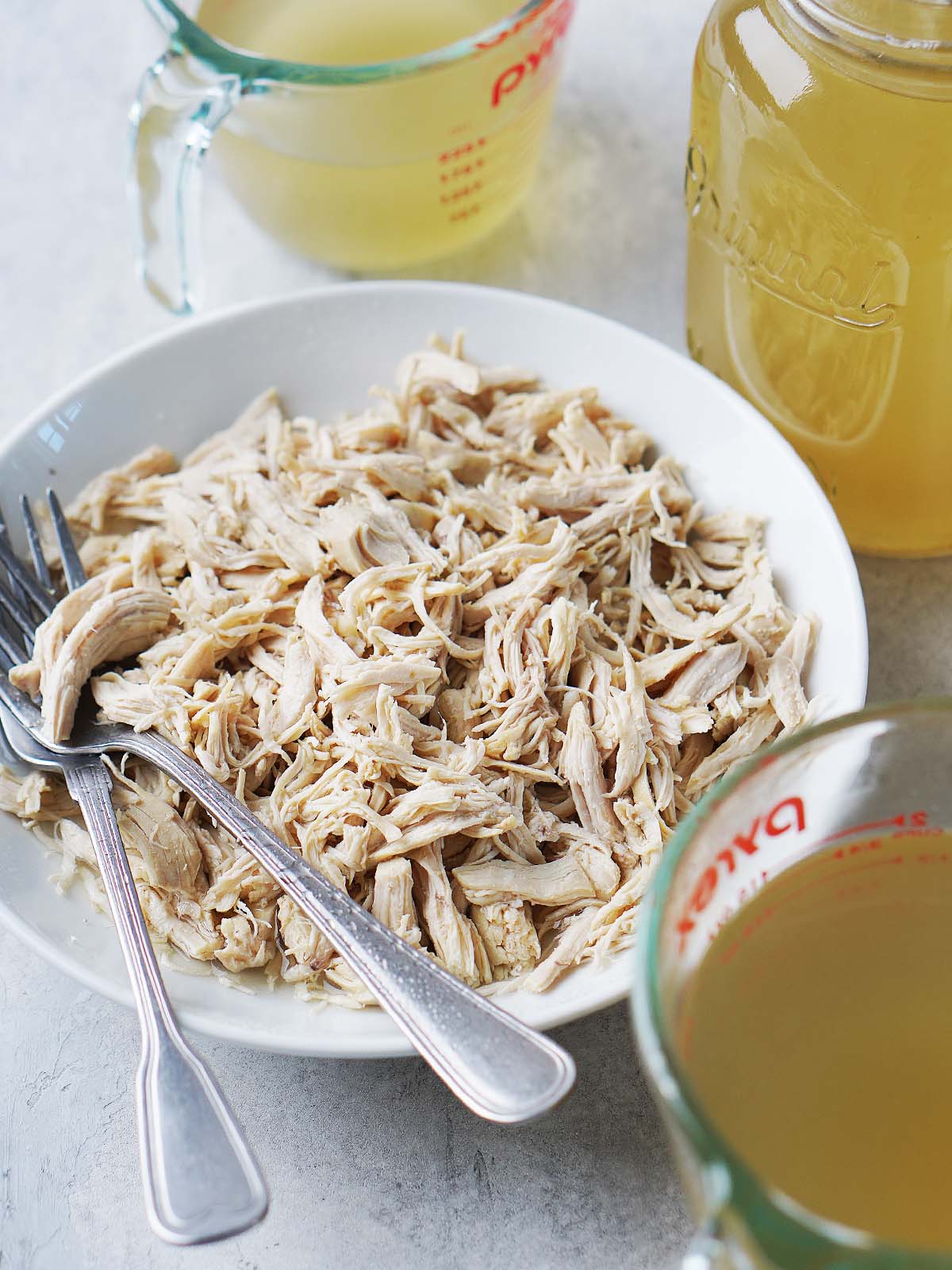 A bowl of shredded cooked chicken with two forks, surrounded by glass measuring cups filled with broth.