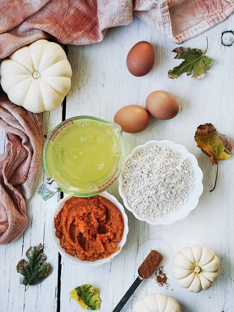 Ingredients for pumpkin baking, including bowls of pumpkin pur&eacute;e and flour, a measuring cup of liquid, three eggs, two small white pumpkins, and scattered dried leaves on a white wooden surface.