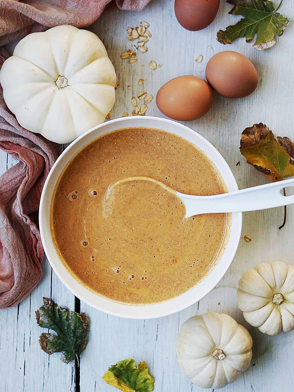 A white bowl of smooth brown batter with a spoon, surrounded by two white pumpkins, two brown eggs, and scattered autumn leaves.