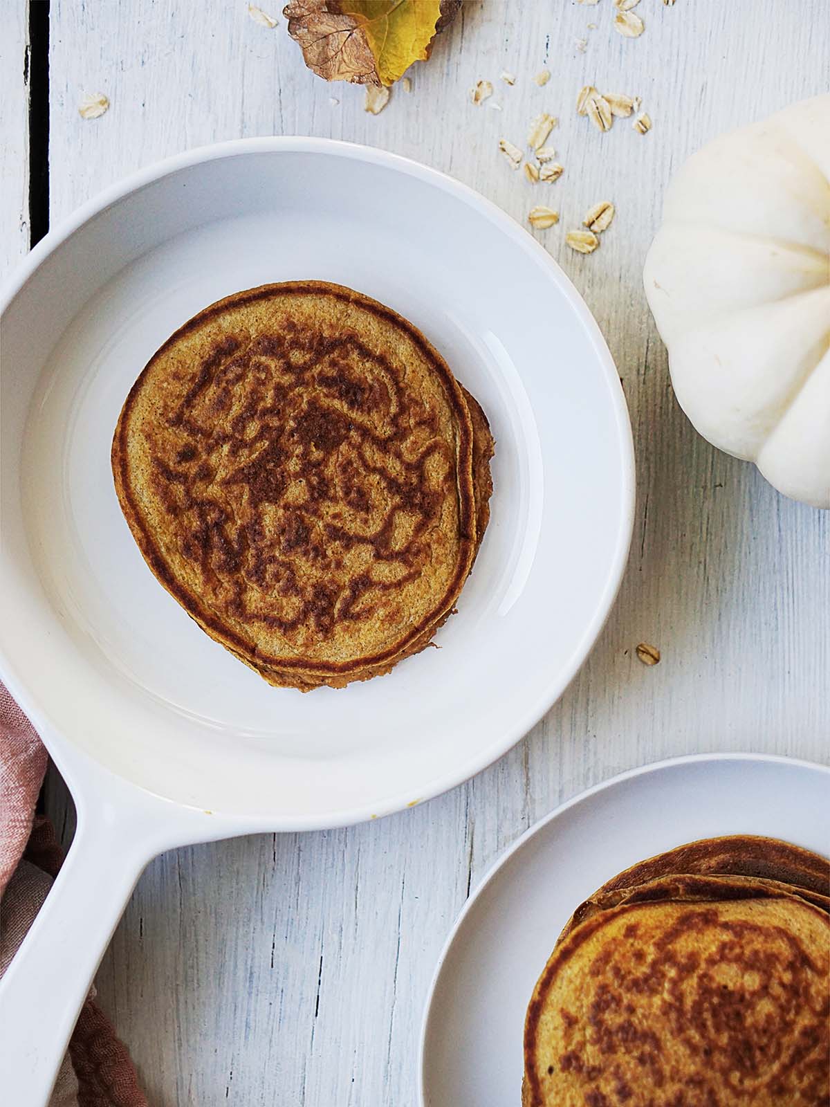 A single pancake in a white skillet with a stack of pancakes on a plate nearby, accompanied by a small white pumpkin.