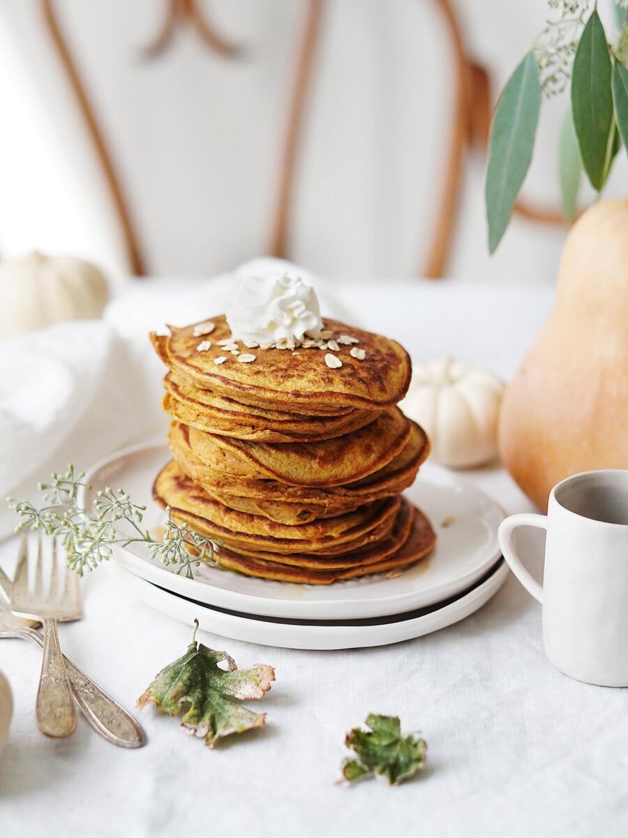 A stack of pumpkin pancakes topped with whipped cream and oats on a white plate.