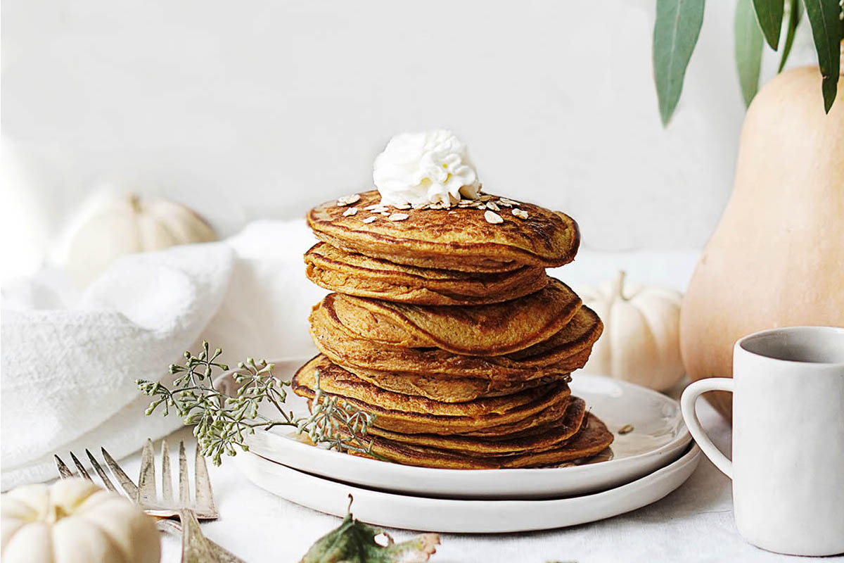 A stack of pumpkin pancakes topped with whipped cream and oats on a plate, with a mug and small pumpkins nearby.