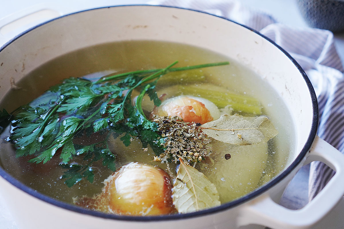 A white pot filled with broth containing onion halves, parsley, and bay leaves.